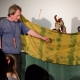 Herec stojí před žlutozelenou plentou, za ní je maňásek kozy. 